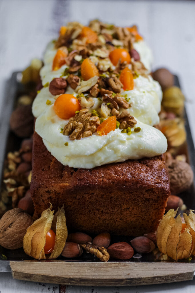 A baked and decorated carrot cake loaf with cream cheese frosting and topping of nuts and fruit.