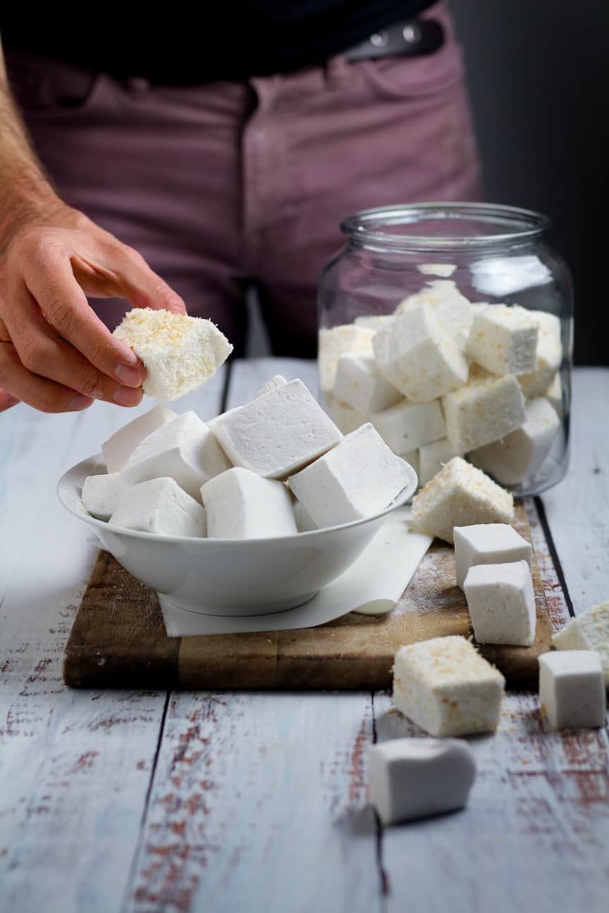 A bowl and jar of homemade marshmallows with the cook's hand holding one marshmallow.