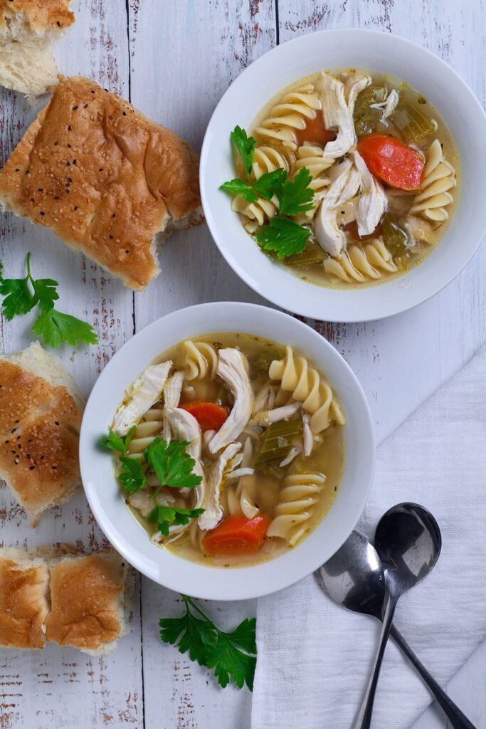 Two bowls of Chicken and Noodle Soup with torn crust bread on a white wooden table.