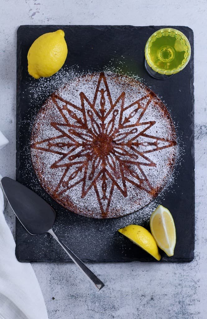 Torta Caprese al limone or Capri Lemon cake on a slate board with lemons, a glass of limoncello and a cake server.