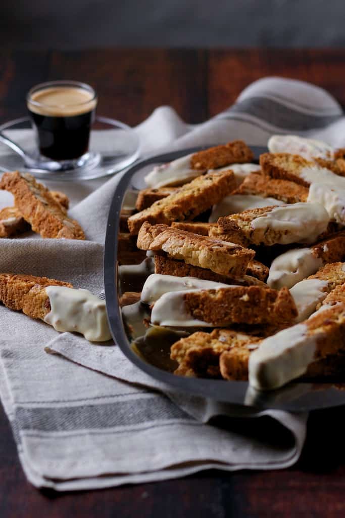 A tray of white chocolate dipped, apricot and almond biscotti on a tray with a small glass of espresso in the background.