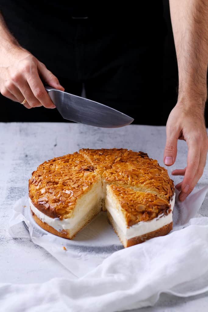 A German Bee Sting Cake on a plate. Hands visible behind the cake holding a knife ready to slice the cake.