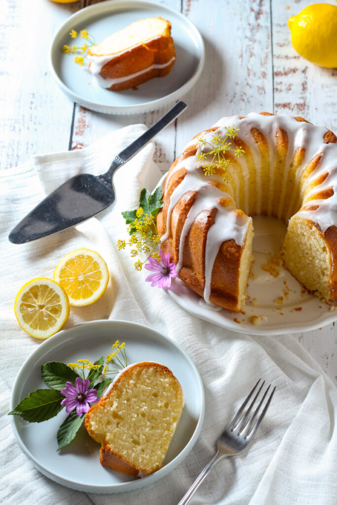 A table with a white cloth, a lemon bundt cake and a plate with a slice of lemon bundt cake.