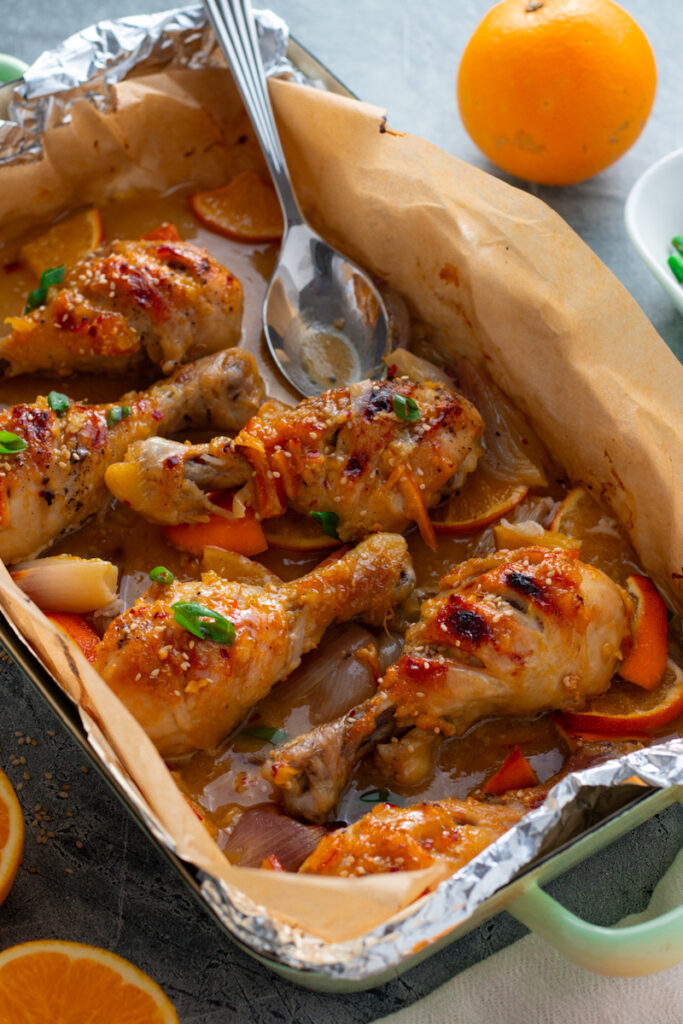 A baking dish with sticky orange chicken drumsticks with fresh oranges and green beans.