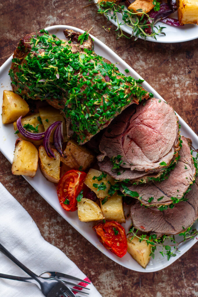Roast Lamb with fresh herb crust, sliced, showing perfect rosy pink, medium-rare interior. Mediterranean Roast vegetables on the side.