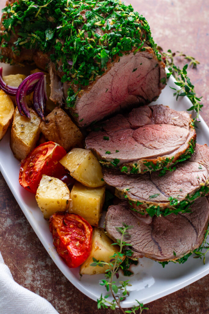 Roast Lamb with fresh herb crust, sliced, showing perfect rosy pink, medium-rare interior. Mediterranean Roast vegetables on the side.