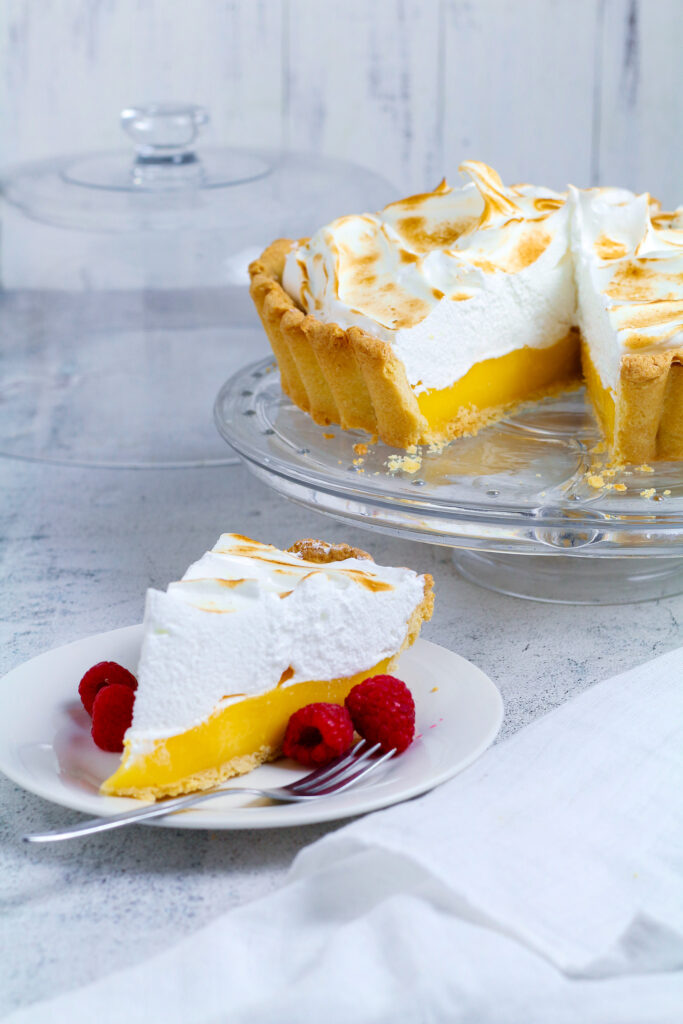 Perfect Lemon Meringue Pie sliced and served with fresh raspberries. Complete pie in the background on a cake stand.