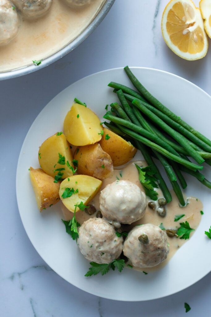 Königsberger Klopse: German meatballs in creamy caper sauce served with potatoes and green beans on a white plate.